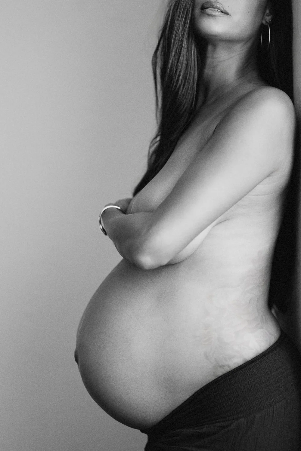 Black and white photo of a pregnant woman with long hair, partially covering her face, arms crossed over her chest, wearing hoop earrings, a bracelet, and black pants.