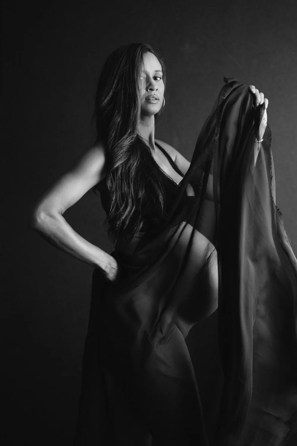 A black and white photograph of a woman with long hair, partially draped in a sheer fabric, looking at the camera with a confident expression, against a dark background.