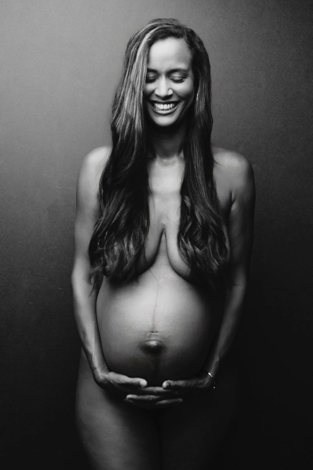 Black-and-white portrait of a pregnant woman with long hair, smiling and holding her belly with both hands, against a plain background.