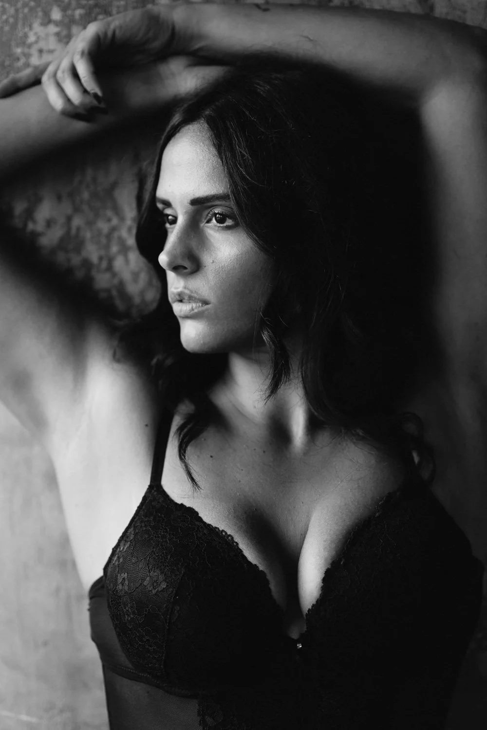 Black and white portrait of a woman with dark hair, wearing a lace bra, lying down with her arm resting on her head, looking away.