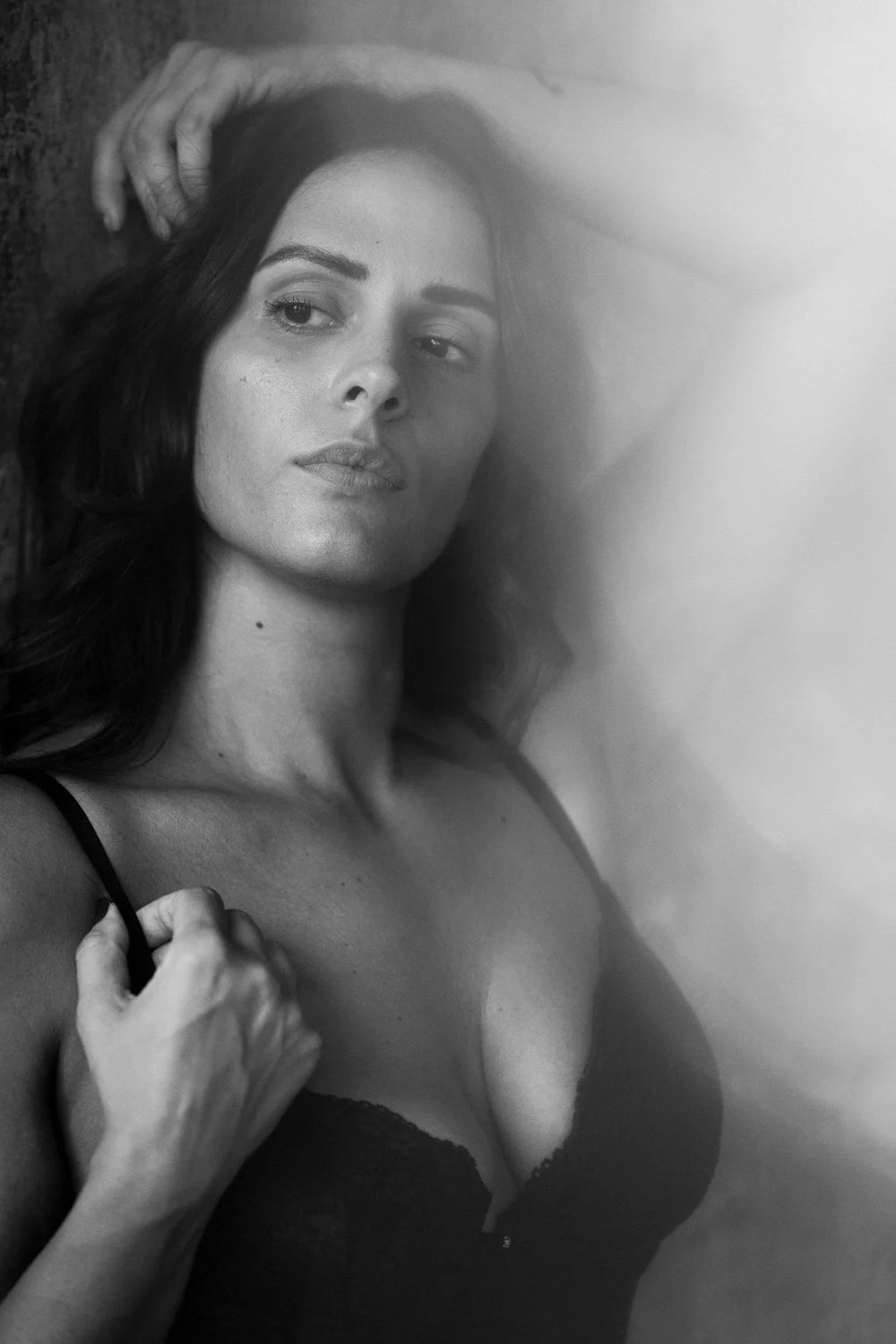 A black-and-white photo of a woman with medium-length hair, lying down with her head resting against a surface. She is wearing a black lace top and looking slightly to the side, with one hand holding her strap near her shoulder.
