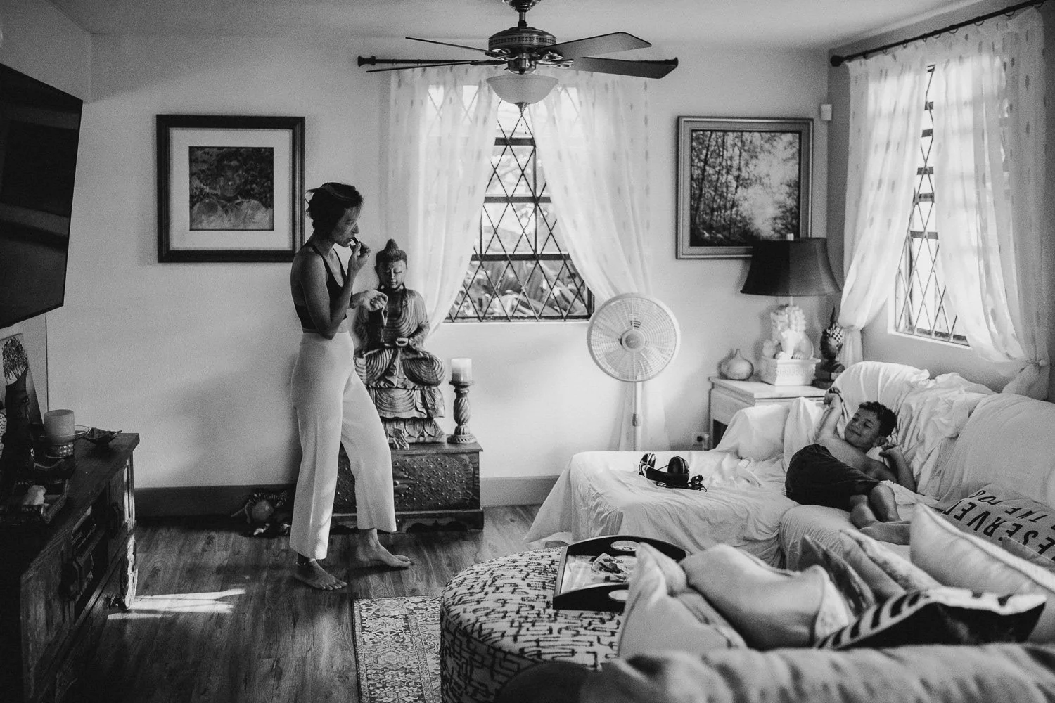 A woman standing and talking to a boy lying on a couch in a living room, with a woman sitting on a small table behind them. The room has curtains, paintings, a fan, and various decorative items.