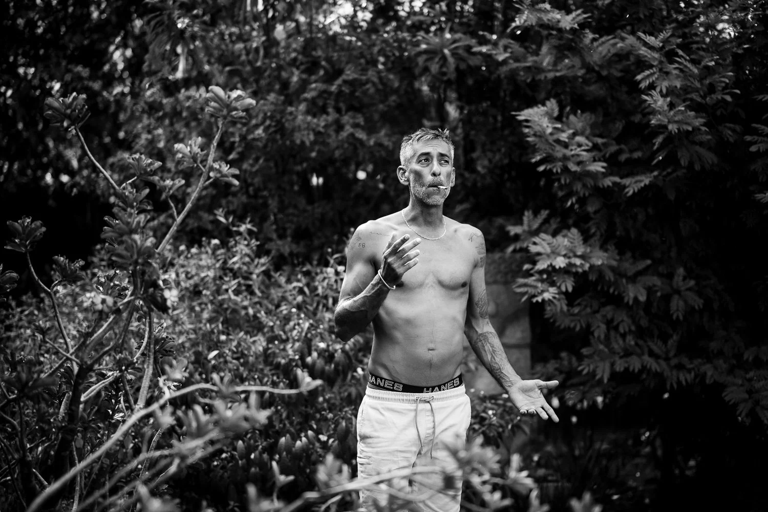 A shirtless man with tattoos around his arms and a necklace, standing outdoors among dense foliage, holding a cigarette in his right hand, with a contemplative expression.