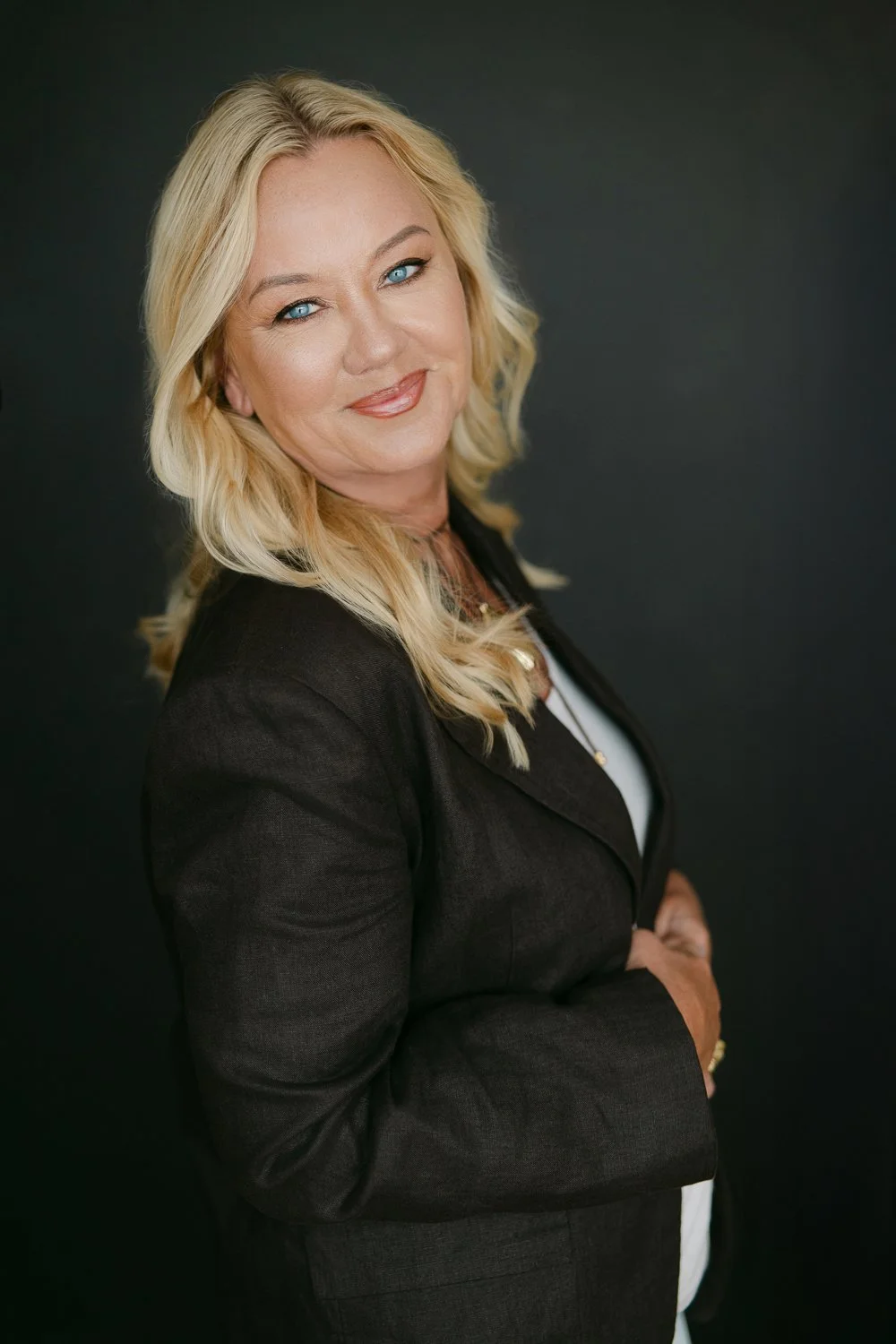 A woman with blonde hair, blue eyes, and light makeup, smiling softly, wearing a black blazer and white top, standing against a dark background.