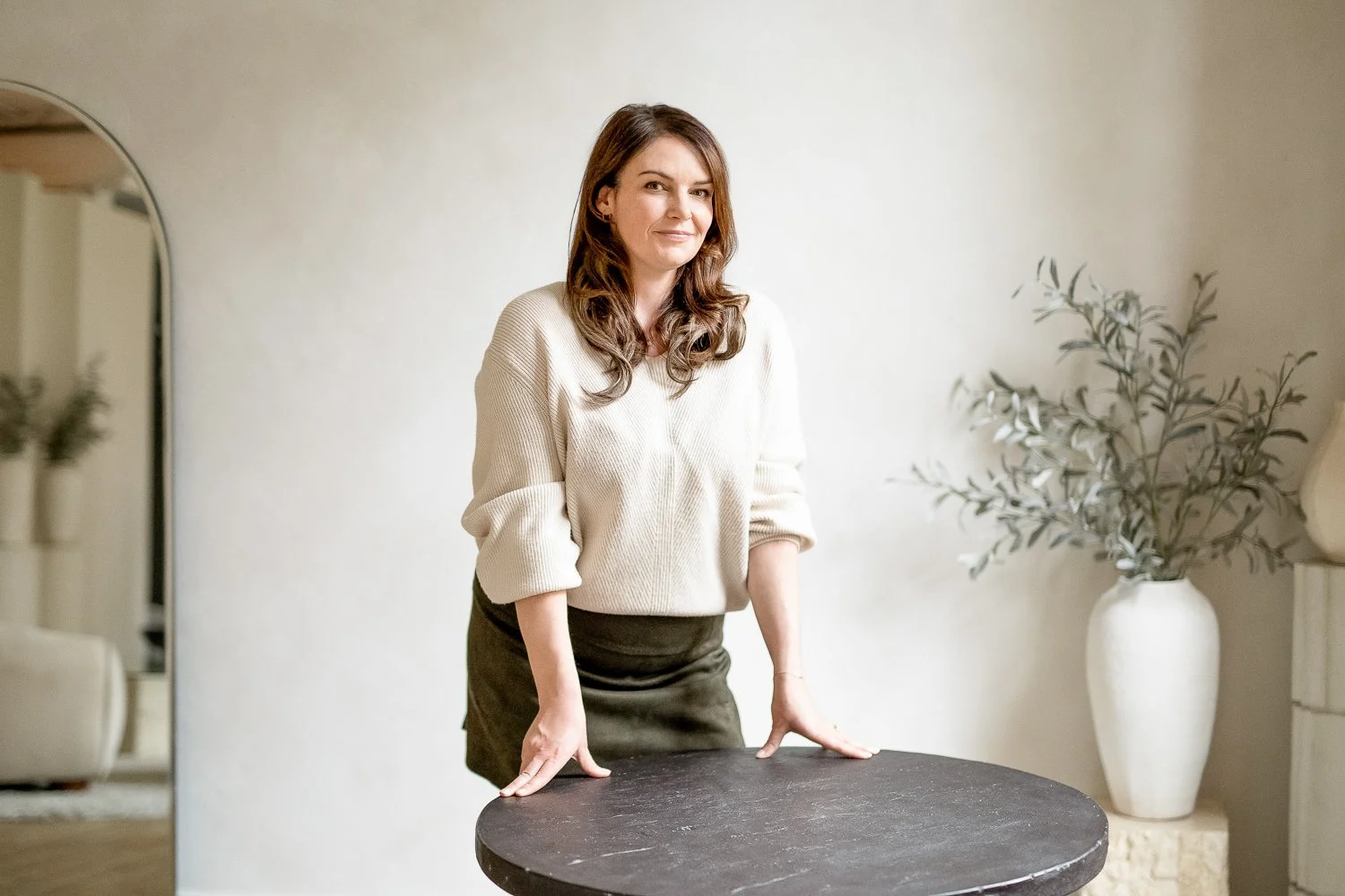 A woman with brown hair smiling, leaning over a round black table in a bright room with beige walls and a large white vase with green leafy branches in the background.