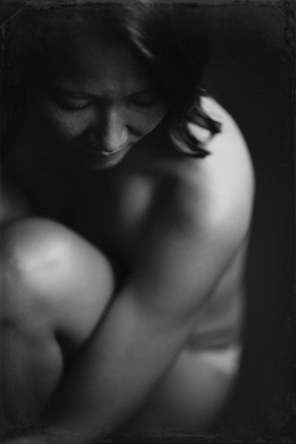 Close-up black and white photograph of a woman with her eyes closed, with her head slightly bowed, highlighting her face and shoulder.