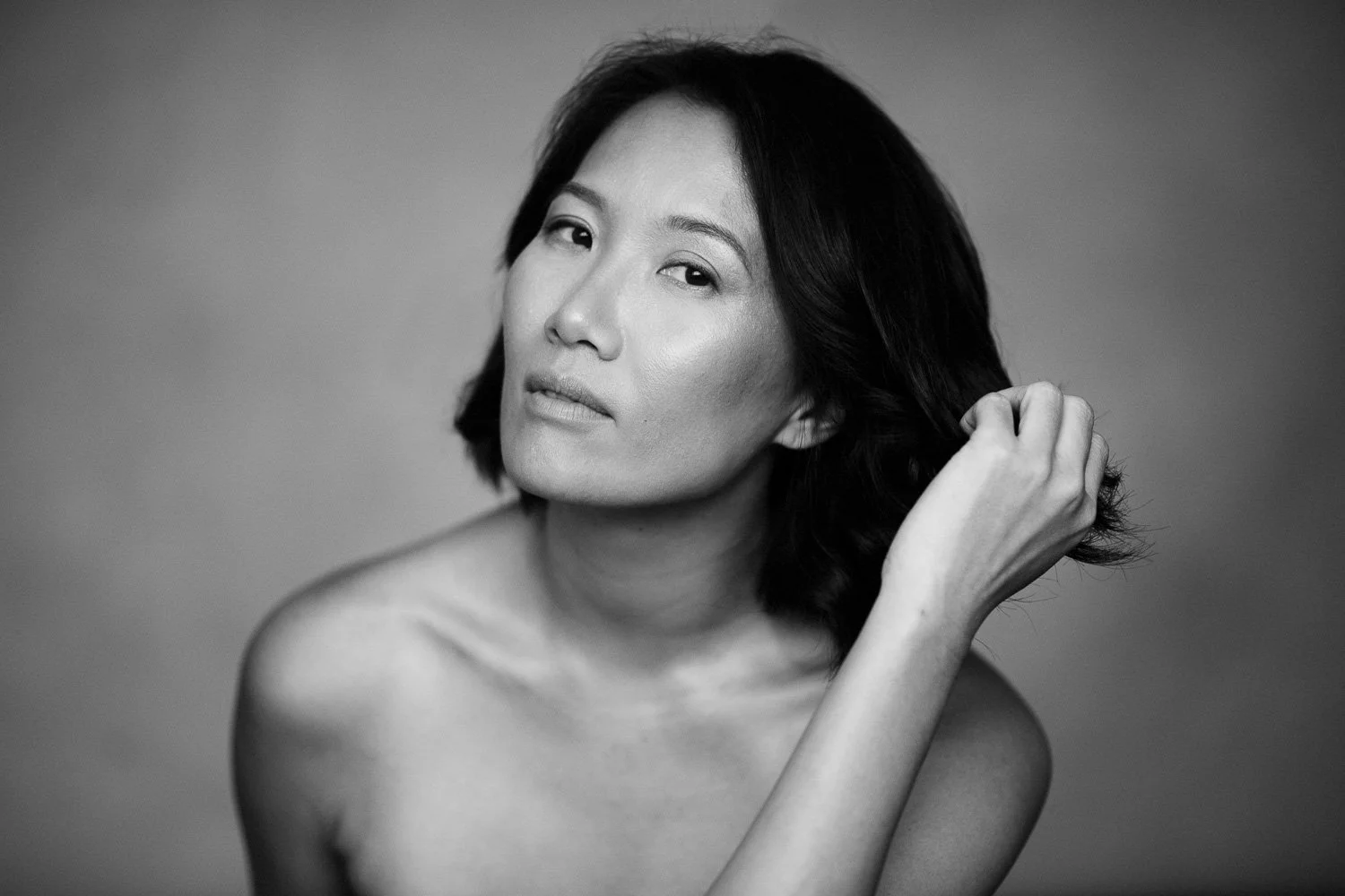 Black and white portrait of a woman with shoulder-length dark hair, gazing at the camera, touching her hair with her right hand, with an expressionless face.