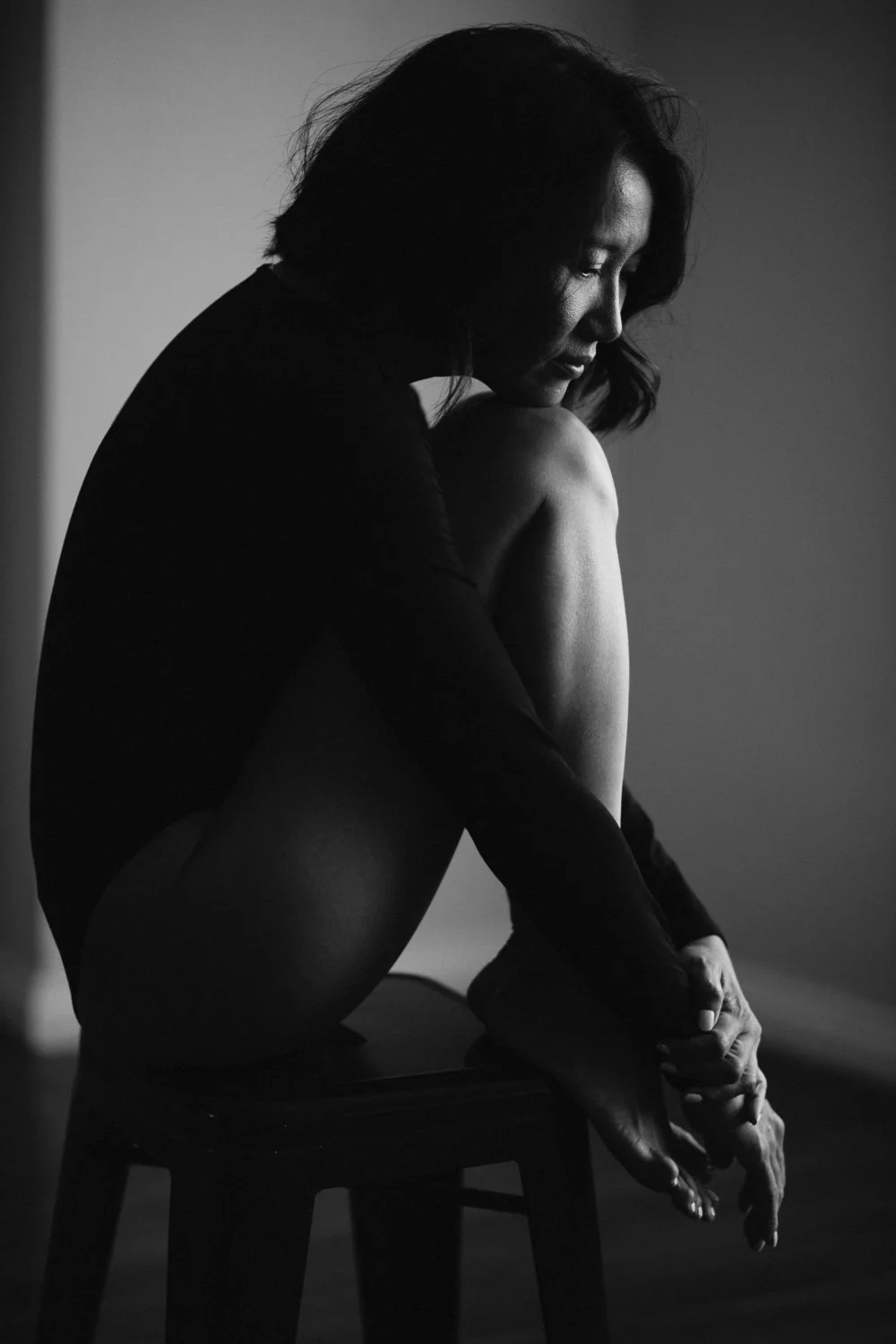 A black and white photo of a woman sitting on a stool with her knee bent and her head resting on her knee. She has shoulder-length dark hair and is wearing a long-sleeve top, with a contemplative or introspective expression.