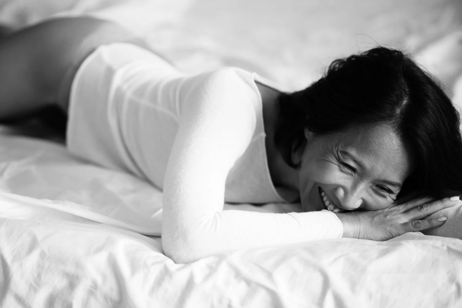 A woman with dark hair lying on a bed, smiling and resting her head on her hands.
