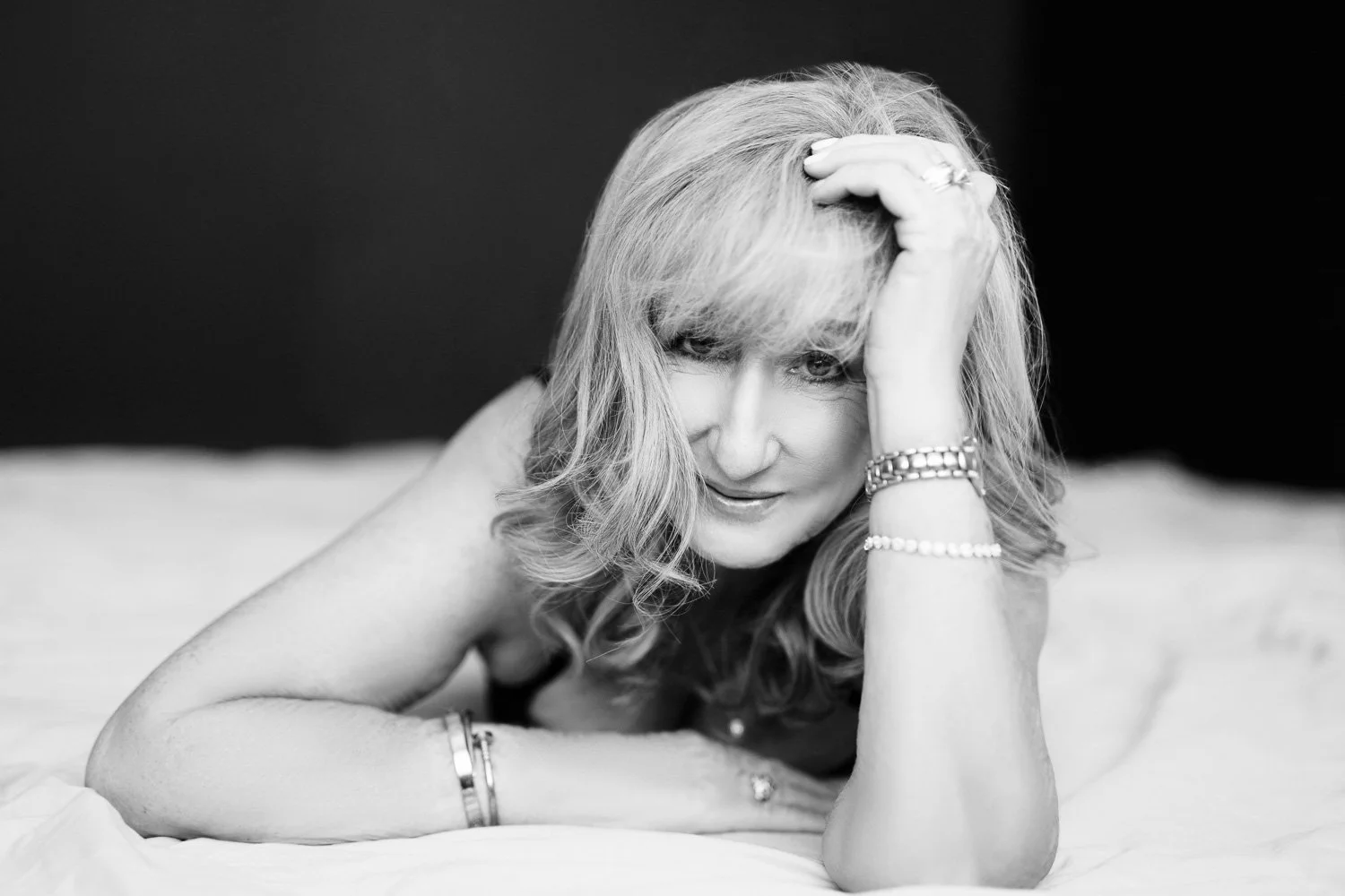 A black and white portrait of a woman with wavy blonde hair, lying on her side on a bed, resting her head on her hand, looking at the camera. She is wearing jewelry, including bracelets on her wrist and rings.