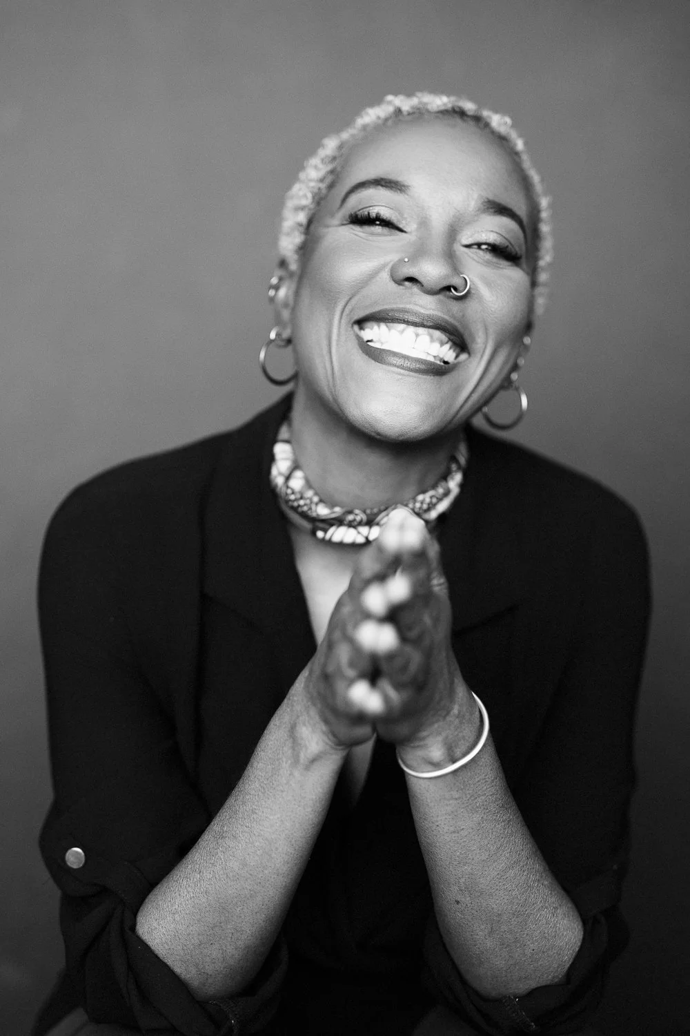 A black and white photo of a smiling woman with short, light-colored hair, wearing hoop earrings, a nose ring, a neck scarf, and a dark jacket. She has her hands clasped together and is looking directly at the camera.