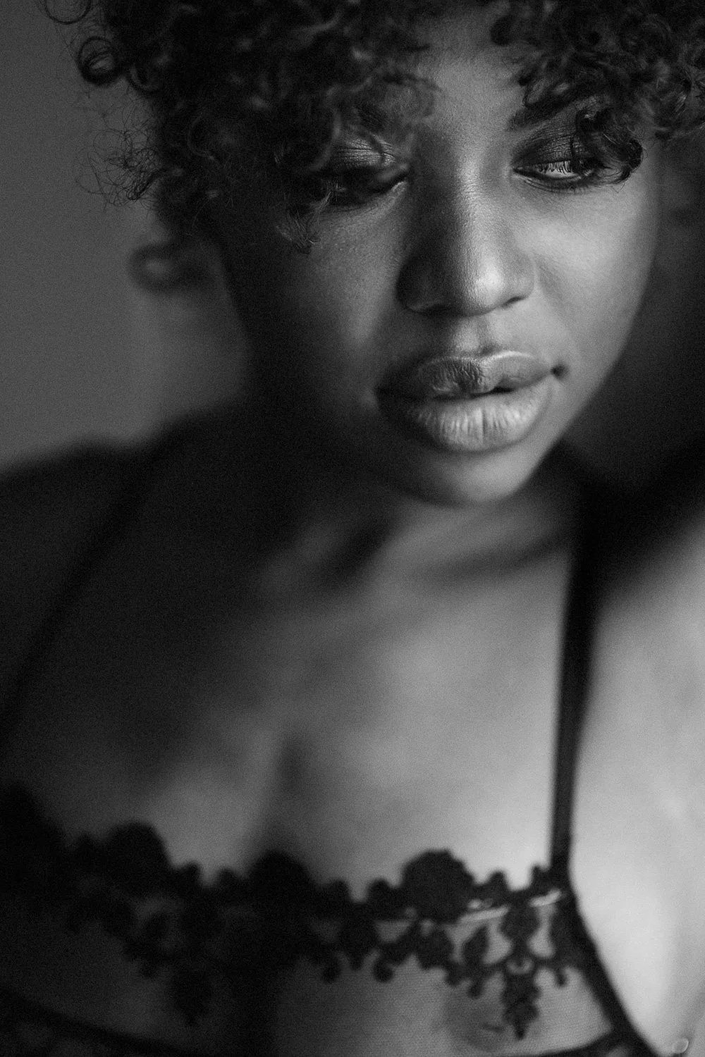 A black-and-white close-up photograph of a woman with curly hair, wearing a lace top, looking slightly down with a pensive expression.
