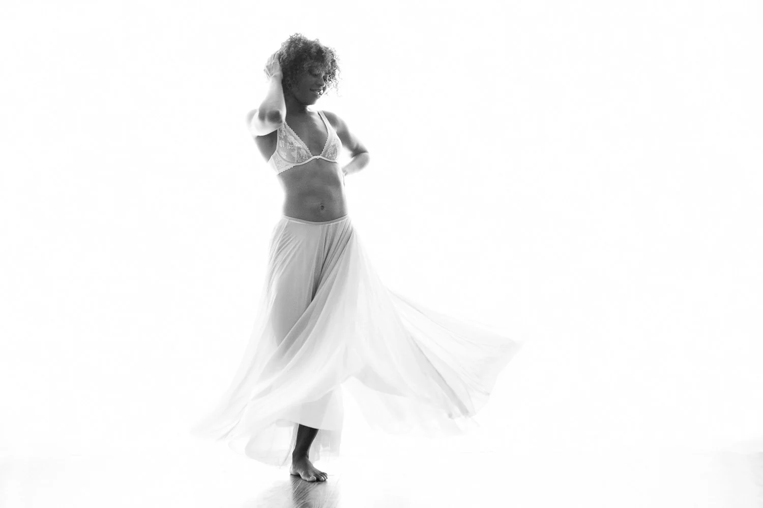 A woman with curly hair in lingerie and a flowing skirt, standing barefoot in a high-key studio shot.