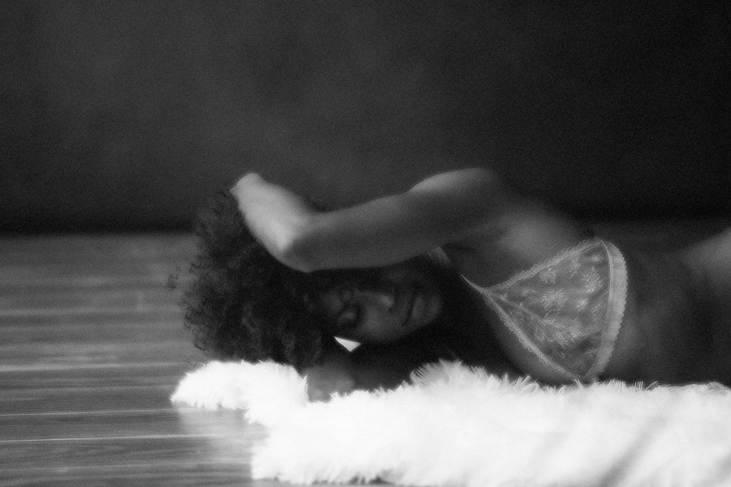 A woman lying on her side on a fluffy surface, with her arm resting on her forehead, wearing a sheer lace top, in black and white photography.