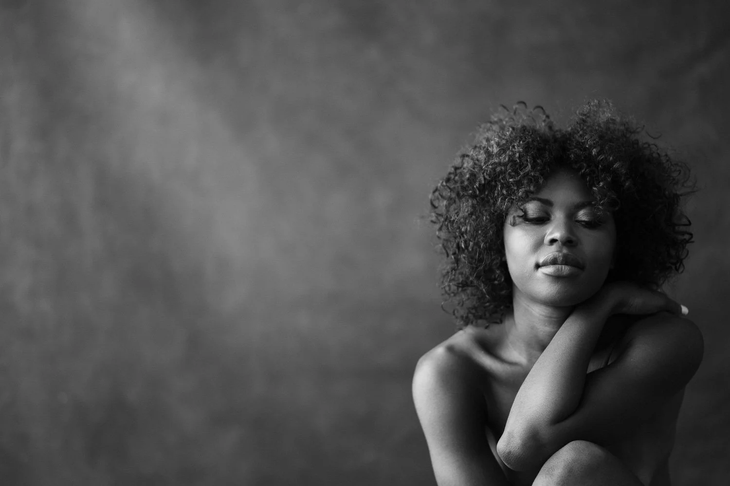 A black and white portrait of a woman with curly hair, her eyes closed, resting her hand on her shoulder, with a textured gray background.