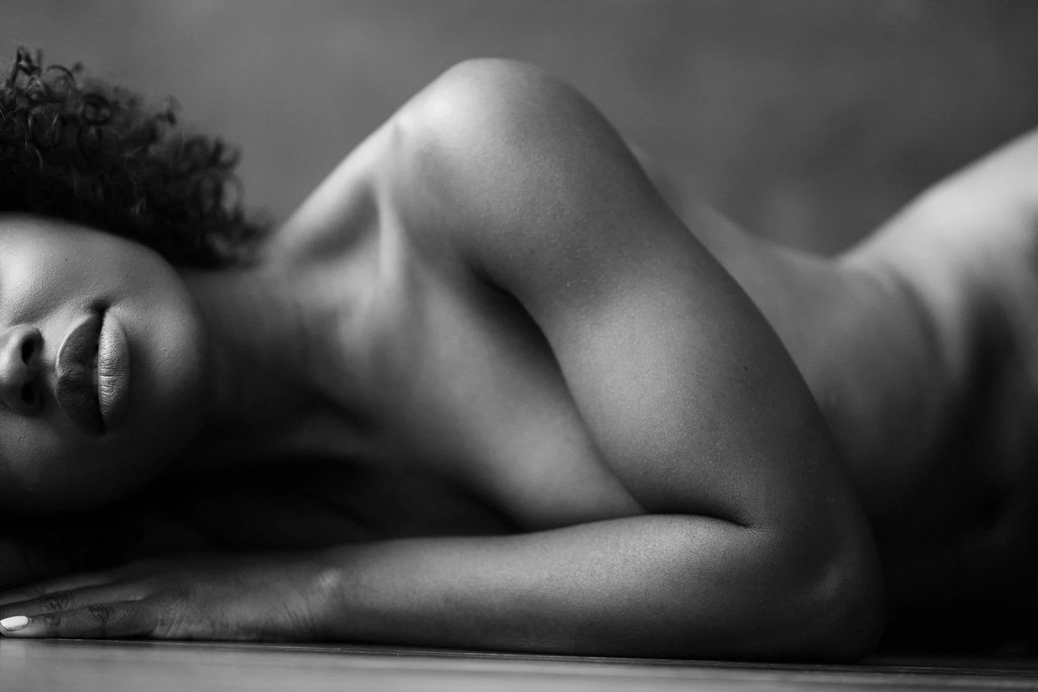 A black and white photo of a woman lying down, with her face resting on her hand, eyes closed, and curly hair visible.