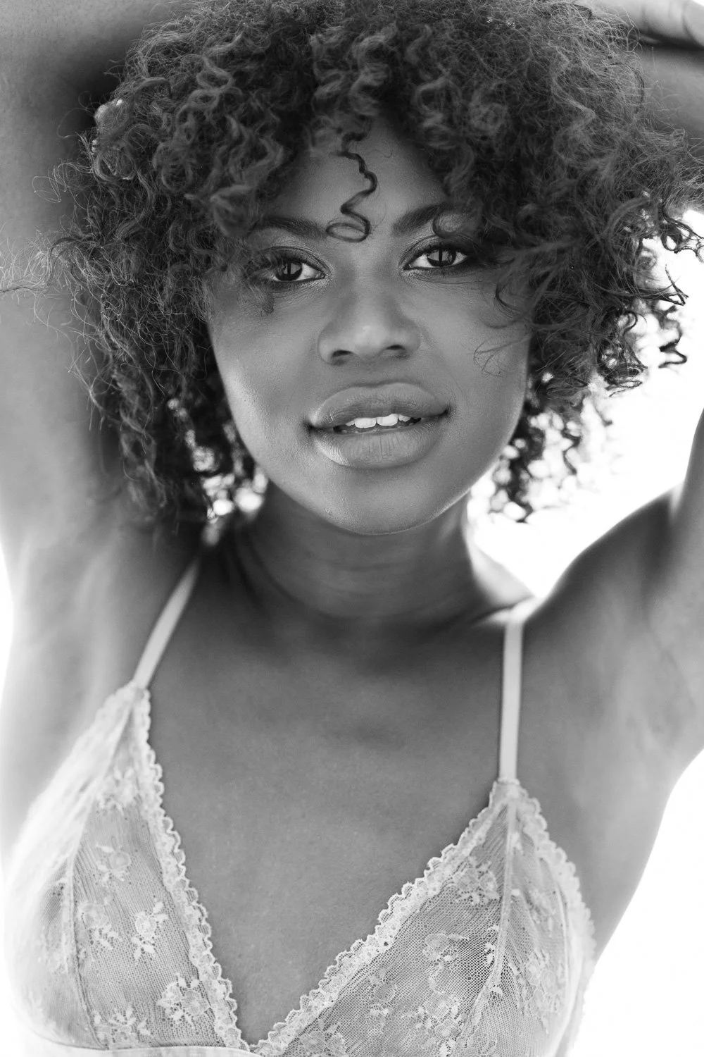 A close-up black and white portrait of a woman with curly hair, wearing a lace top, looking directly into the camera with her arms raised behind her head.