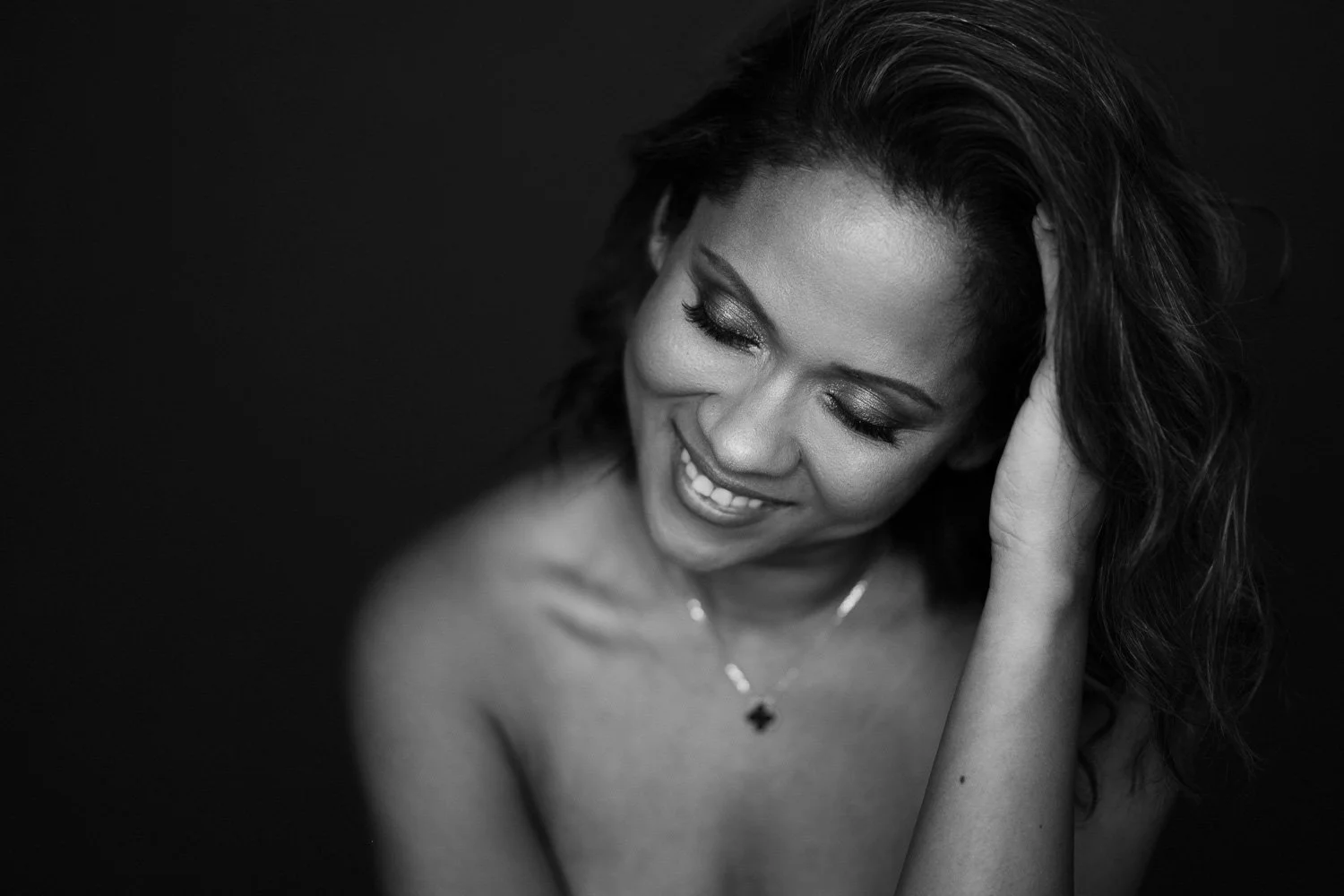 Black and white portrait of a woman smiling with eyes closed, head tilted, and hand touching her hair, wearing a necklace with a small pendant.