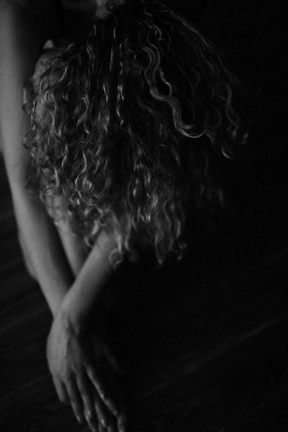 A black and white photo of a person with curly hair, head bowed down, sitting with hands resting on a dark surface.