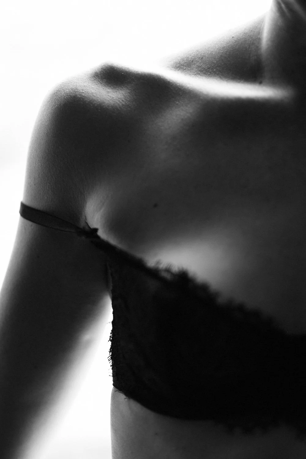 Close-up black and white photo of a woman's bare shoulder, collarbone, and part of black lace bra strap.