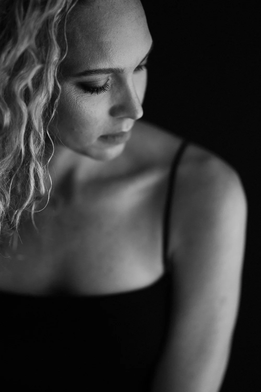 Black and white close-up of a woman with curly hair, wearing a spaghetti strap top, looking downward with eyes closed.