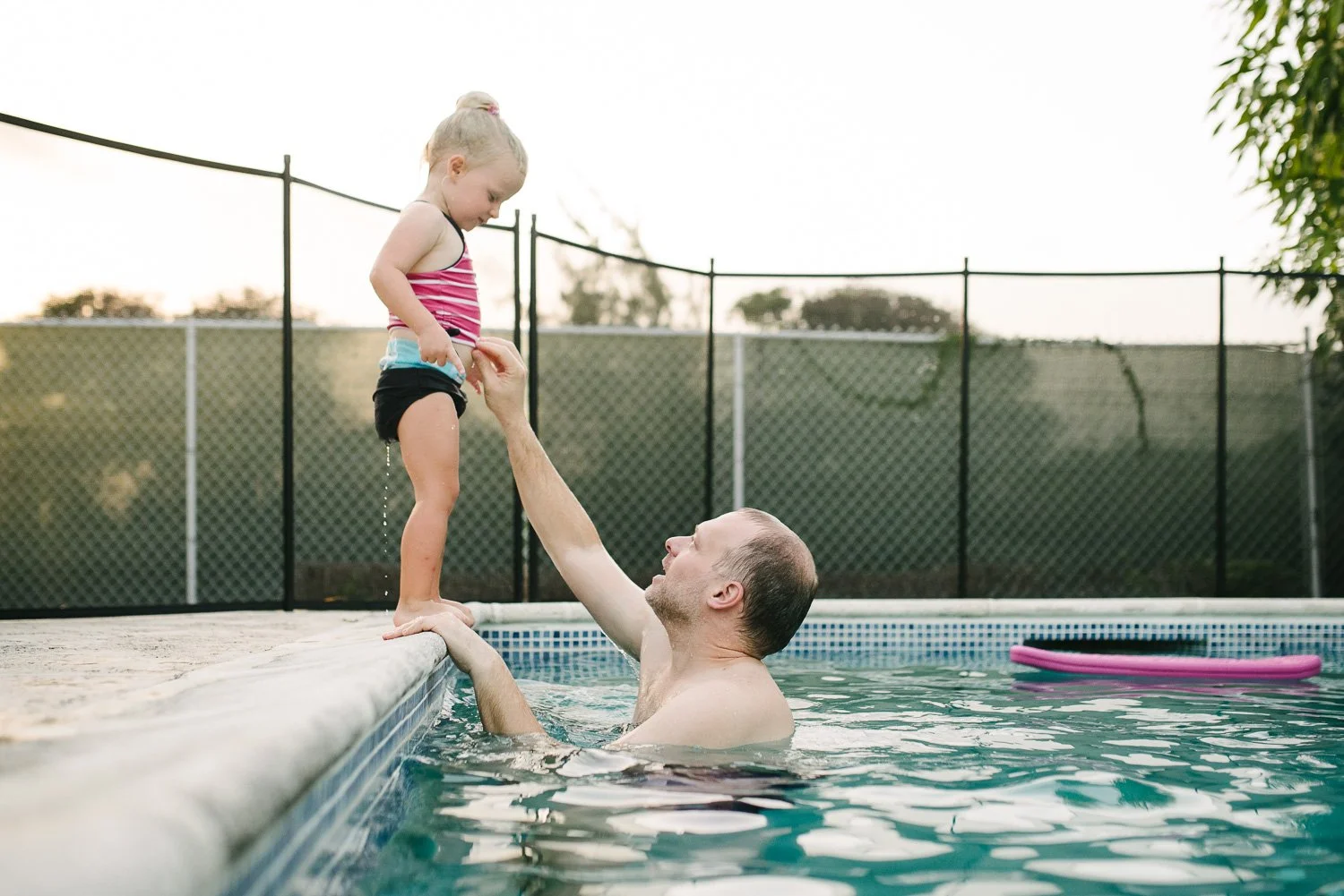 A man is in a swimming pool holding a young girl by the hands, helping her stand at the pool's edge. The girl is looking down at her swimsuit while standing on the pool ledge. The scene is outdoors during late afternoon or early evening with trees an