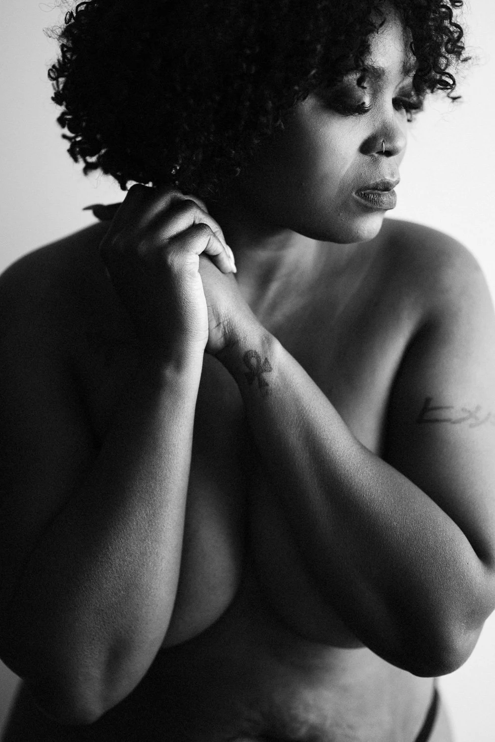 A black and white photograph of a woman with curly hair, looking downward with her eyes closed, holding her hands near her face, and showing tattoos on her arm.