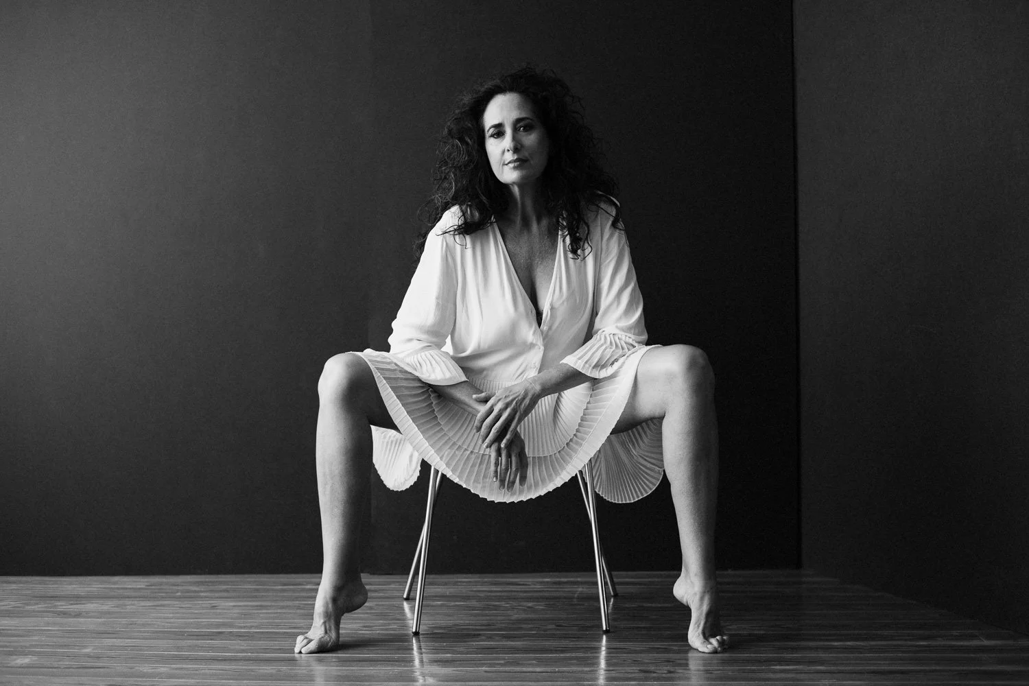 Black and white photo of a woman sitting on a chair with legs wide apart, hands resting on her knees, wearing a loose white blouse and a pleated skirt, in a minimalistic room with dark wall and hardwood floor.