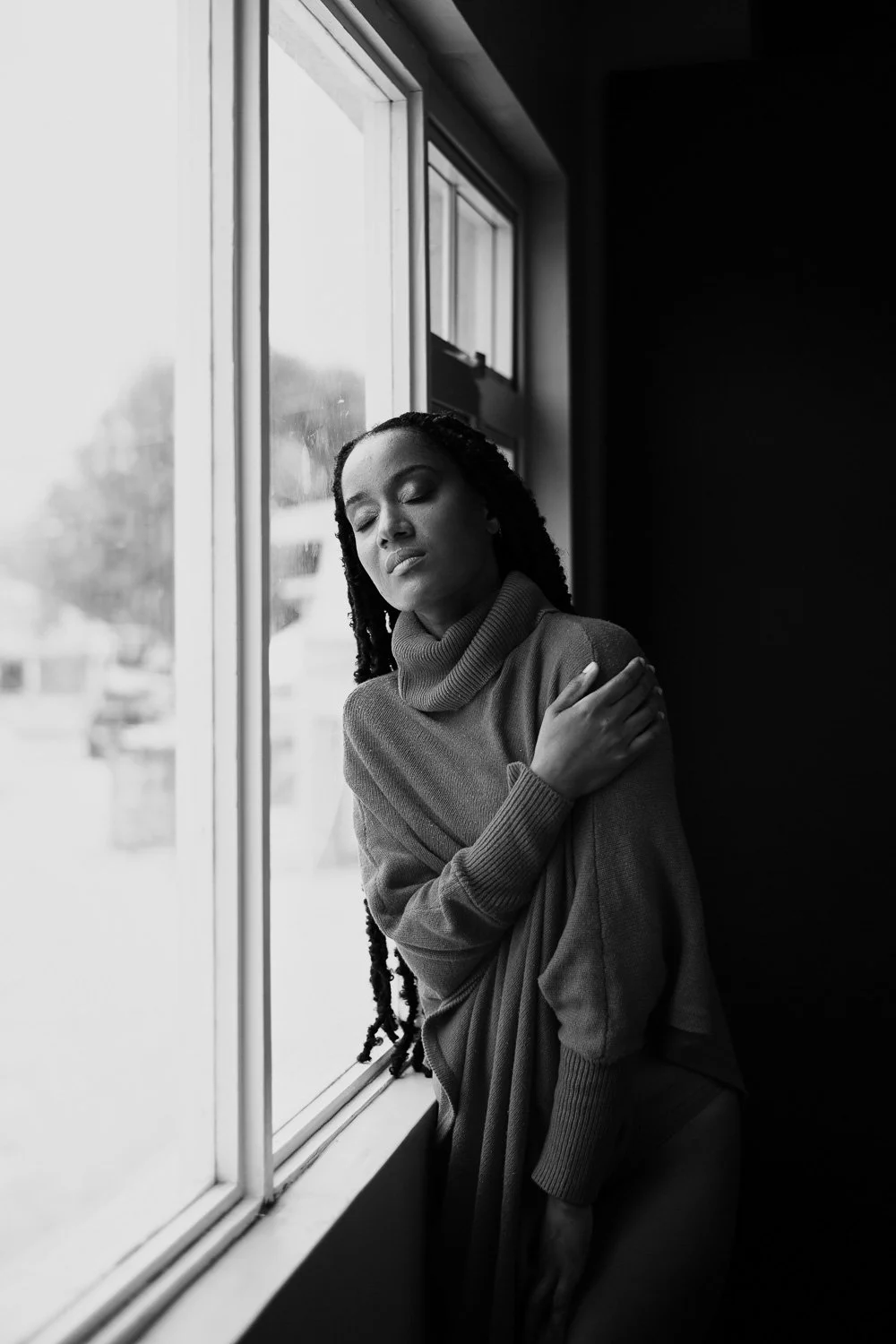A woman with closed eyes and a pained expression, standing by a window with her hand on her shoulder, in black and white.