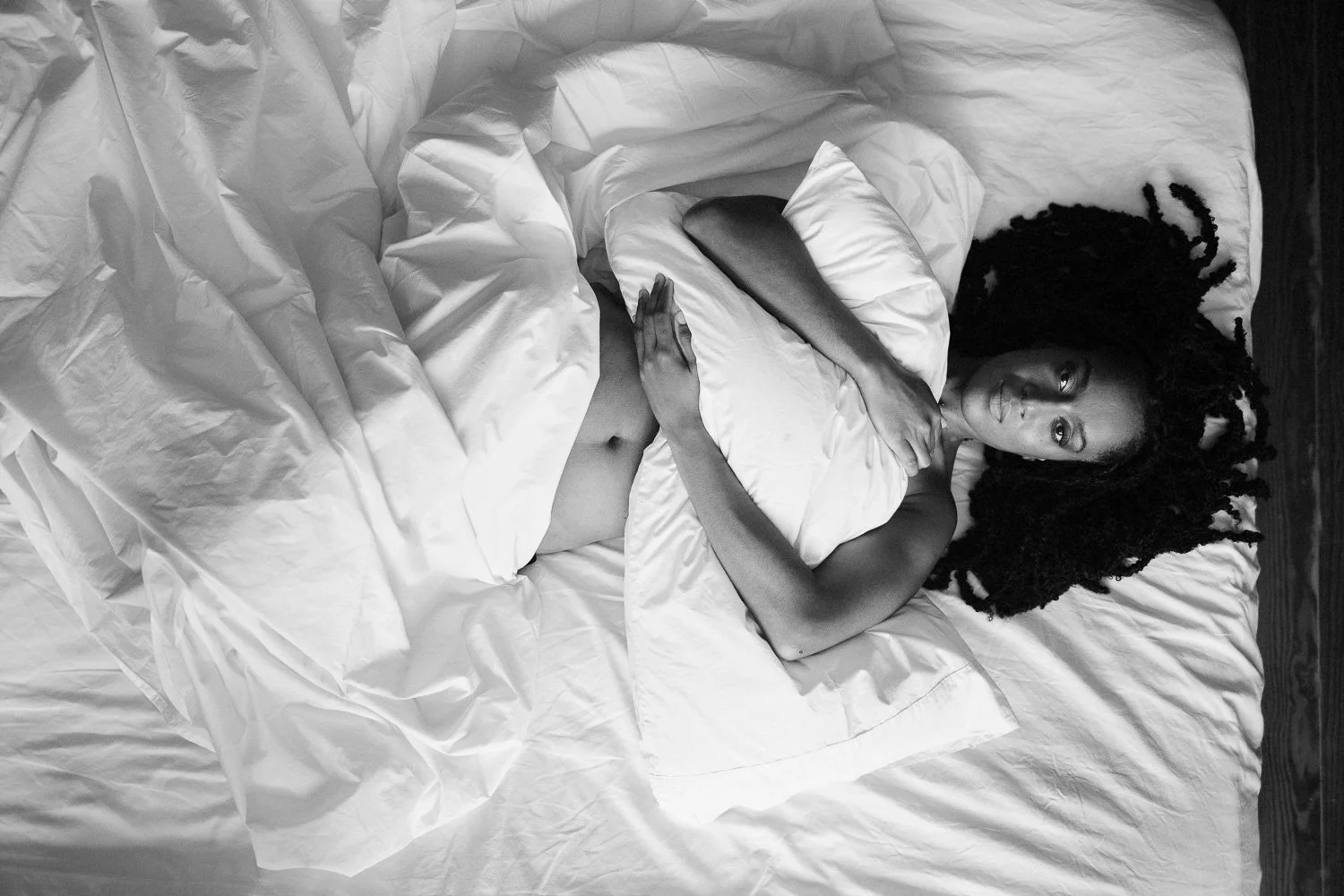 A woman with long dreadlocks reclining in bed, holding a pillow, with her eyes directed at the camera. The bed has white sheets and pillows, and the photo is in black and white.