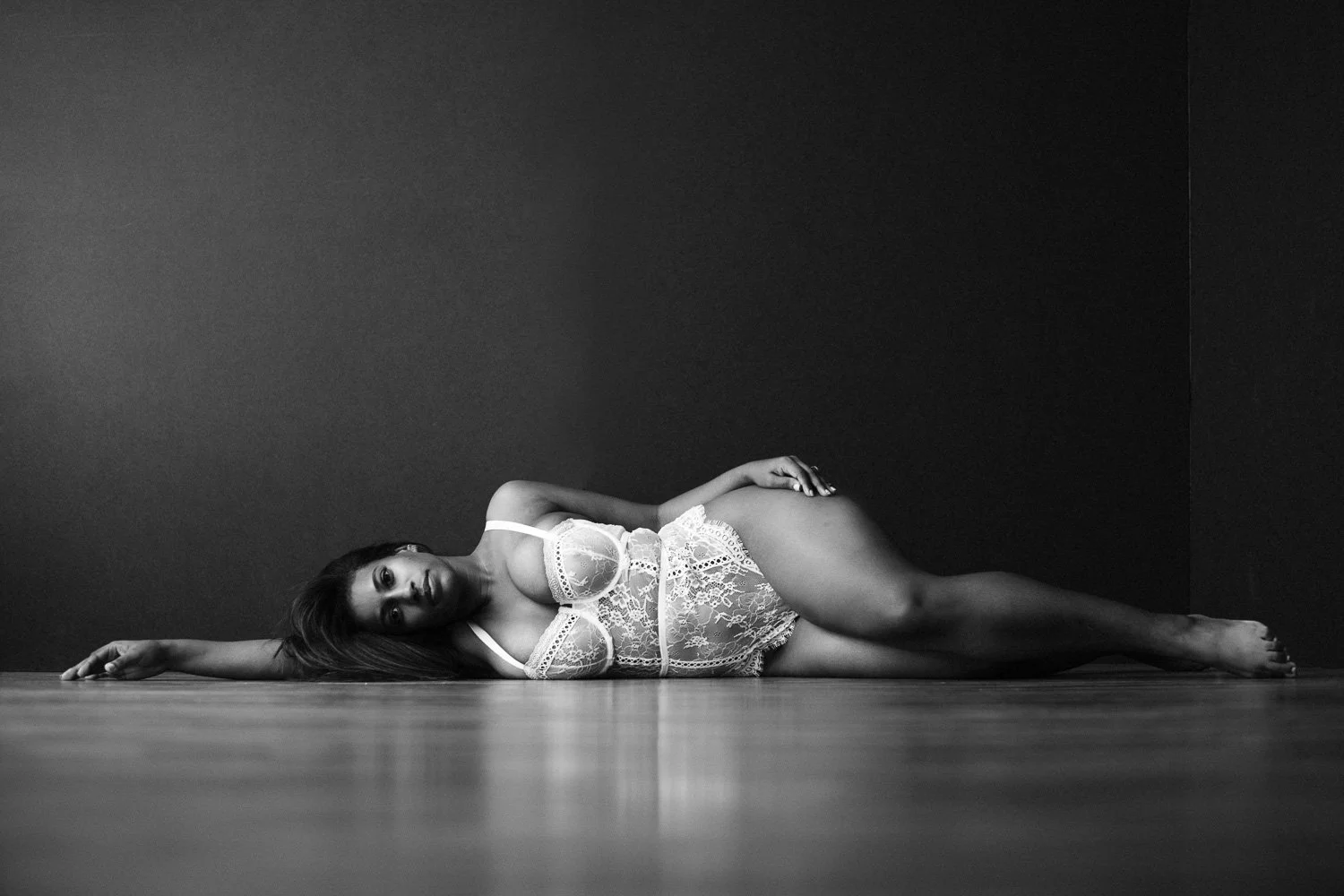 Black and white photo of a woman lying on the floor, wearing lace lingerie, with a dark wall in the background.