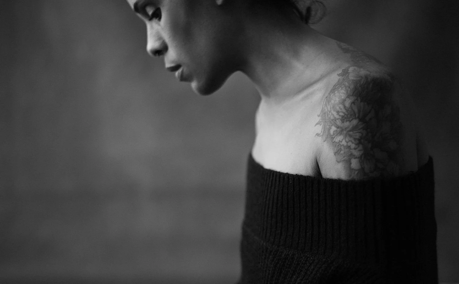 A black and white photo of a woman with short hair, side-profile, wearing an off-the-shoulder sweater, and showing a tattoo on her shoulder.