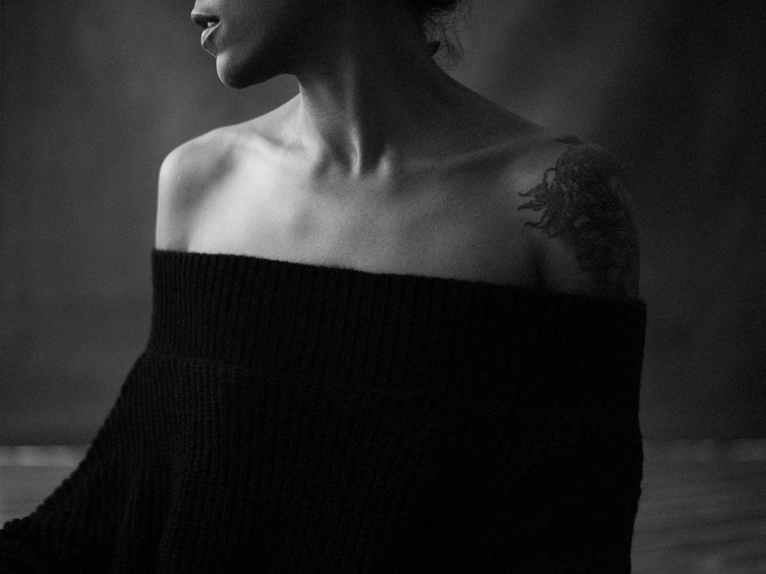 Black and white photo of a woman with short hair, wearing an off-the-shoulder sweater, showing her tattooed shoulder and neck, facing slightly to the left.
