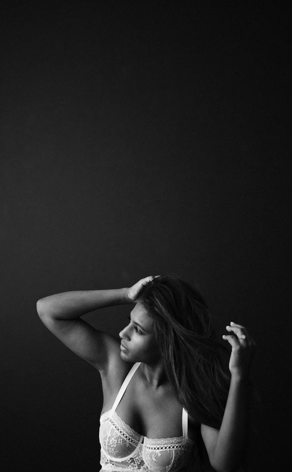 A woman with long hair wearing a lace top, posing with her hand in her hair against a dark background.