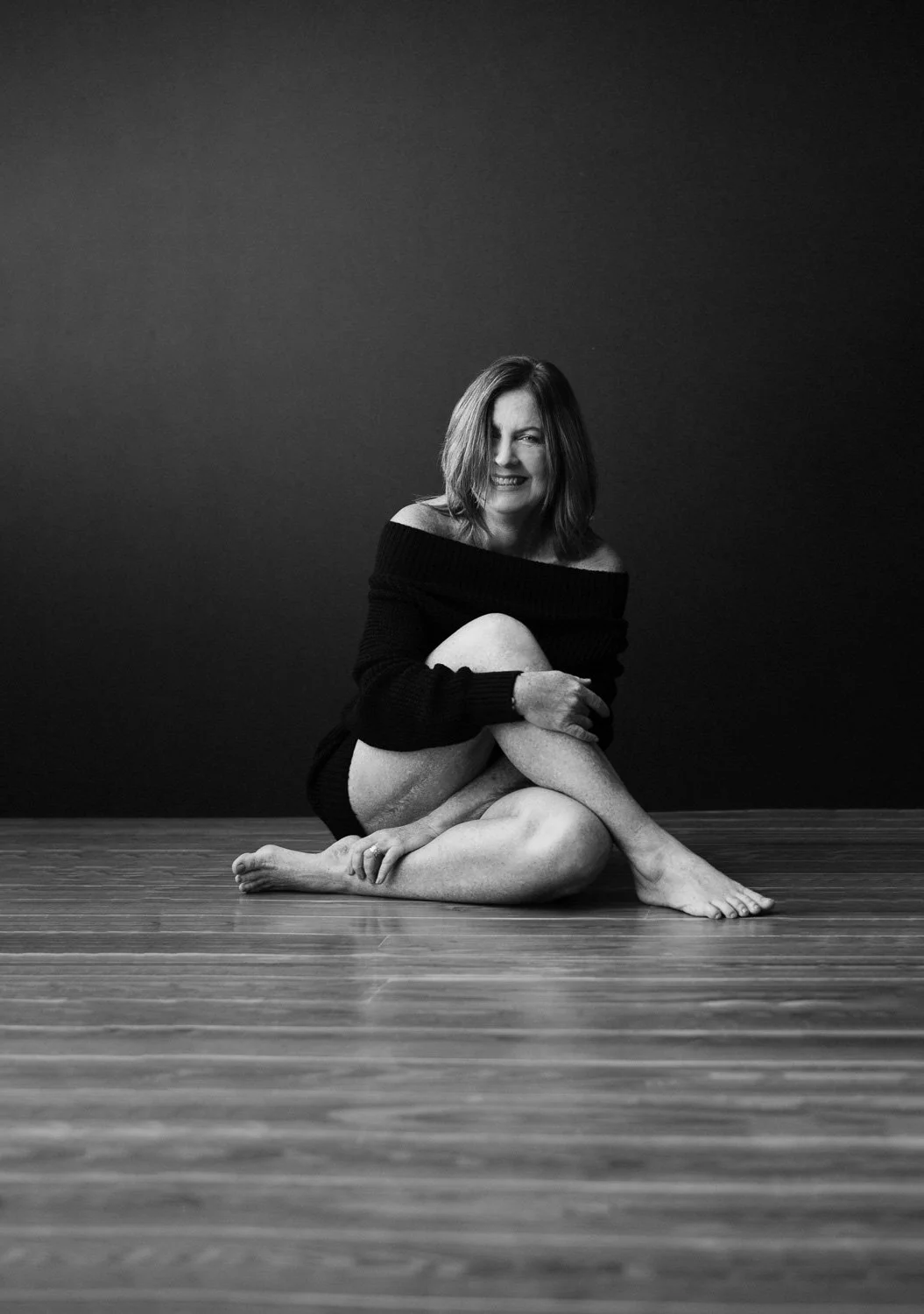 A woman with shoulder-length hair, wearing an off-the-shoulder black sweater, sitting barefoot on a wooden floor against a dark background. She is smiling and holding one of her legs, which is crossed over the other.