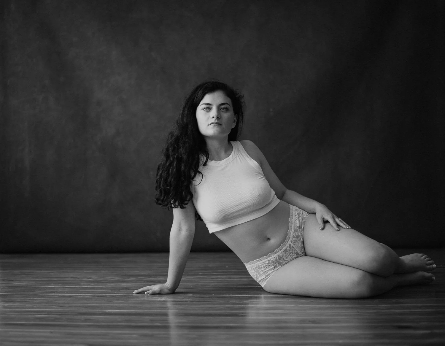 Black and white photo of a woman with long dark hair, wearing a sleeveless top and lace shorts, sitting on a wooden floor with a plain dark background.