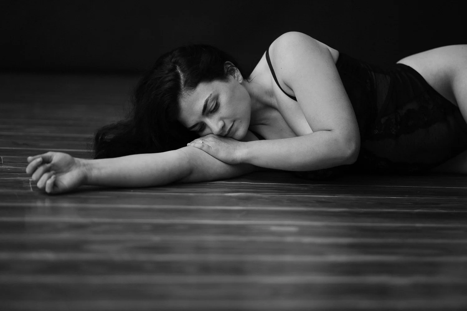 A woman lying on a wooden floor, resting her head and arm on the surface, with her eyes closed, dressed in black lace clothing.