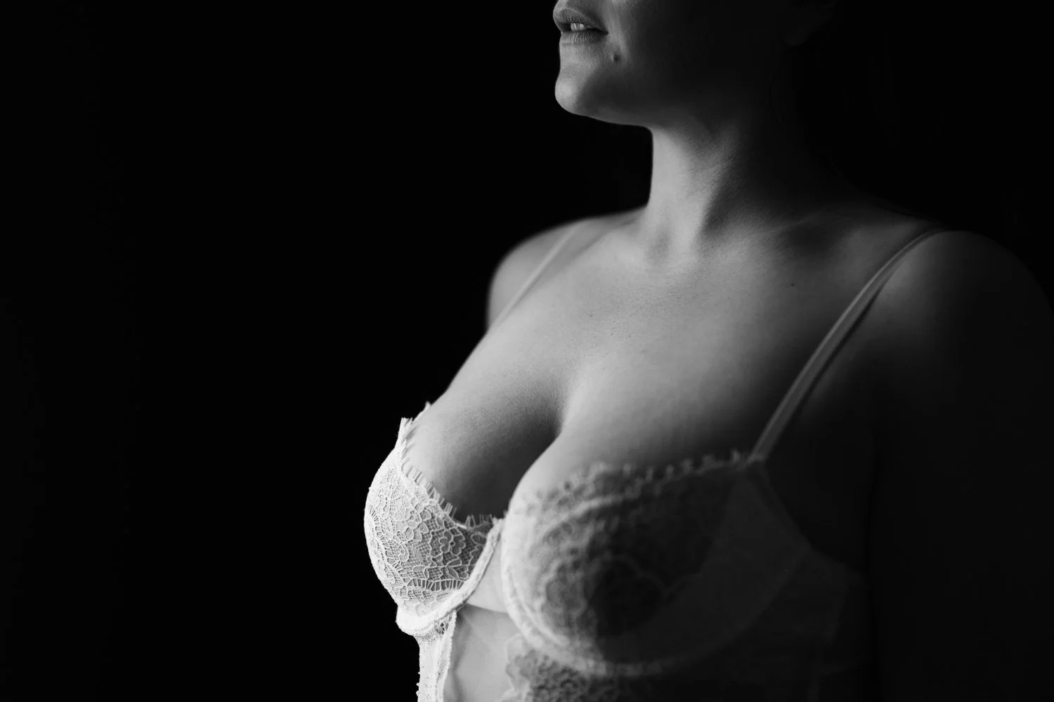 Close-up black and white photo of a woman's face and upper body wearing lace lingerie, with dramatic lighting on her neck and chest.