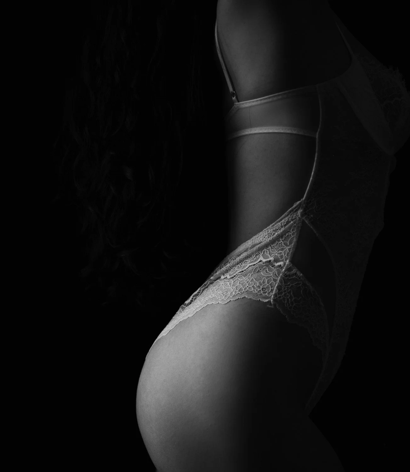Close-up black and white photo of a woman's back and shoulder, wearing a lace bra and lingerie with straps.