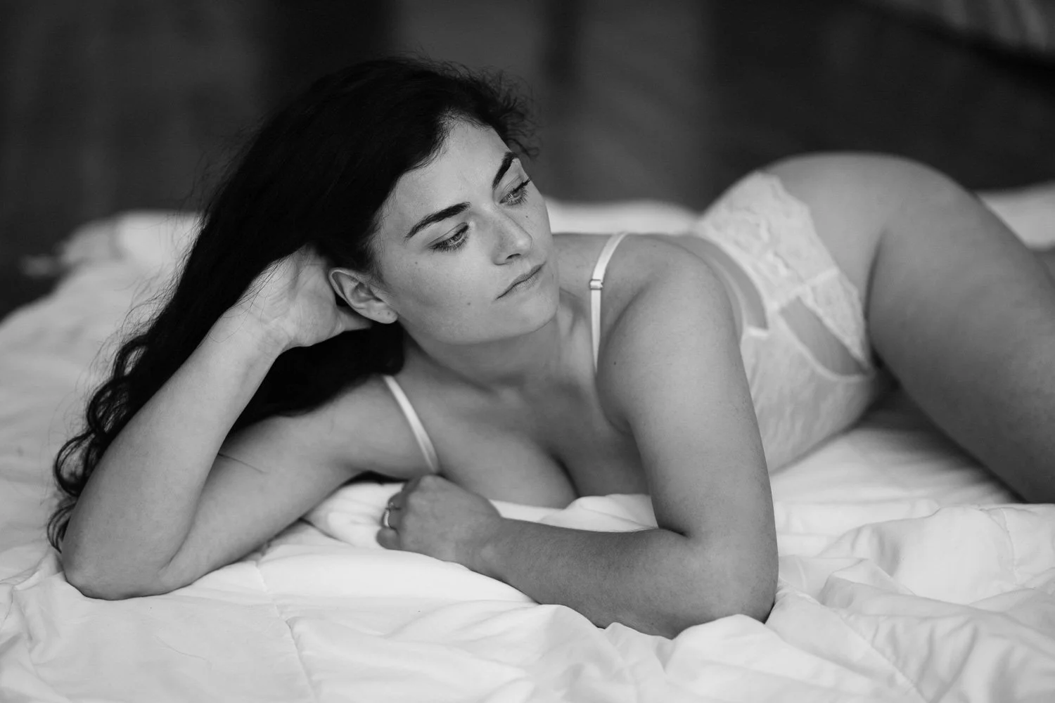 A woman lying on her side on a bed, with dark hair, wearing lingerie, looking off to the side in a relaxed pose.