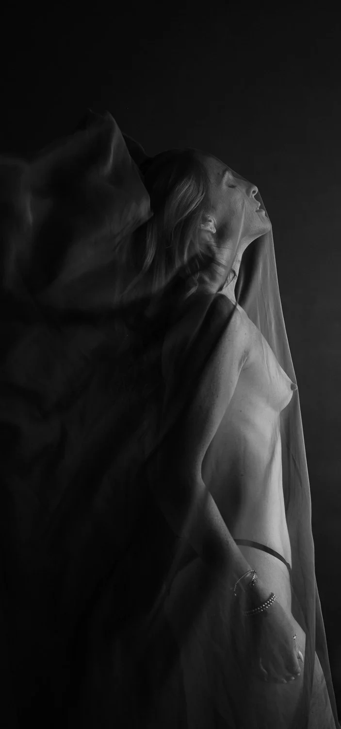 A black and white photograph of a woman with closed eyes, wearing a sheer veil and jewelry, with her head tilted back and a contemplative expression.
