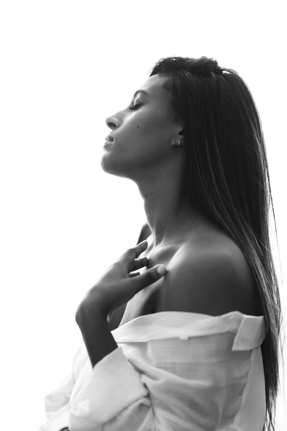 A black-and-white photograph of a woman with closed eyes and long hair, wearing an off-shoulder shirt with one shoulder exposed, touching her collarbone with one hand.
