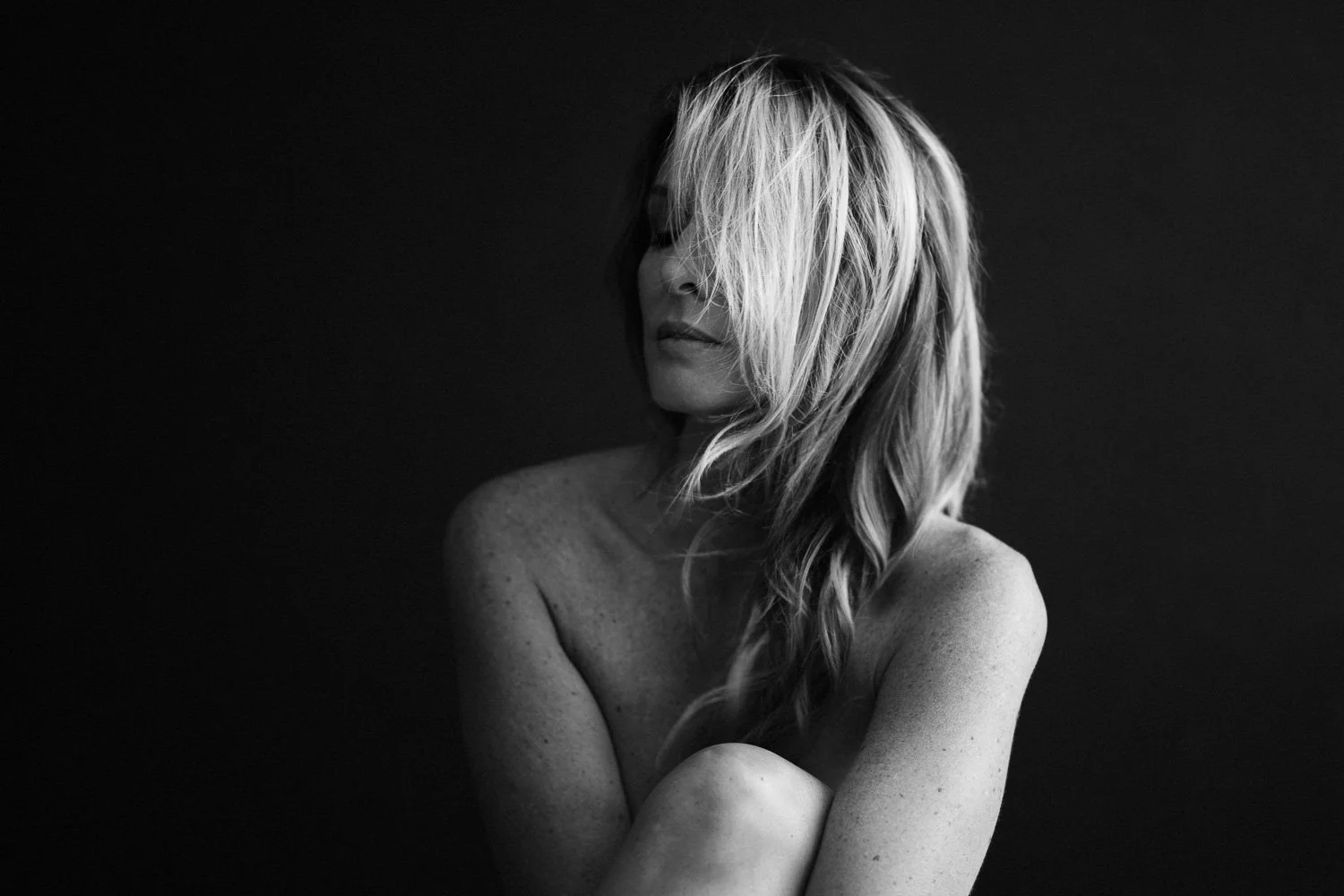 Black and white photo of a woman with long, wavy hair covering part of her face, sitting with her arms crossed against her knees, against a dark background.