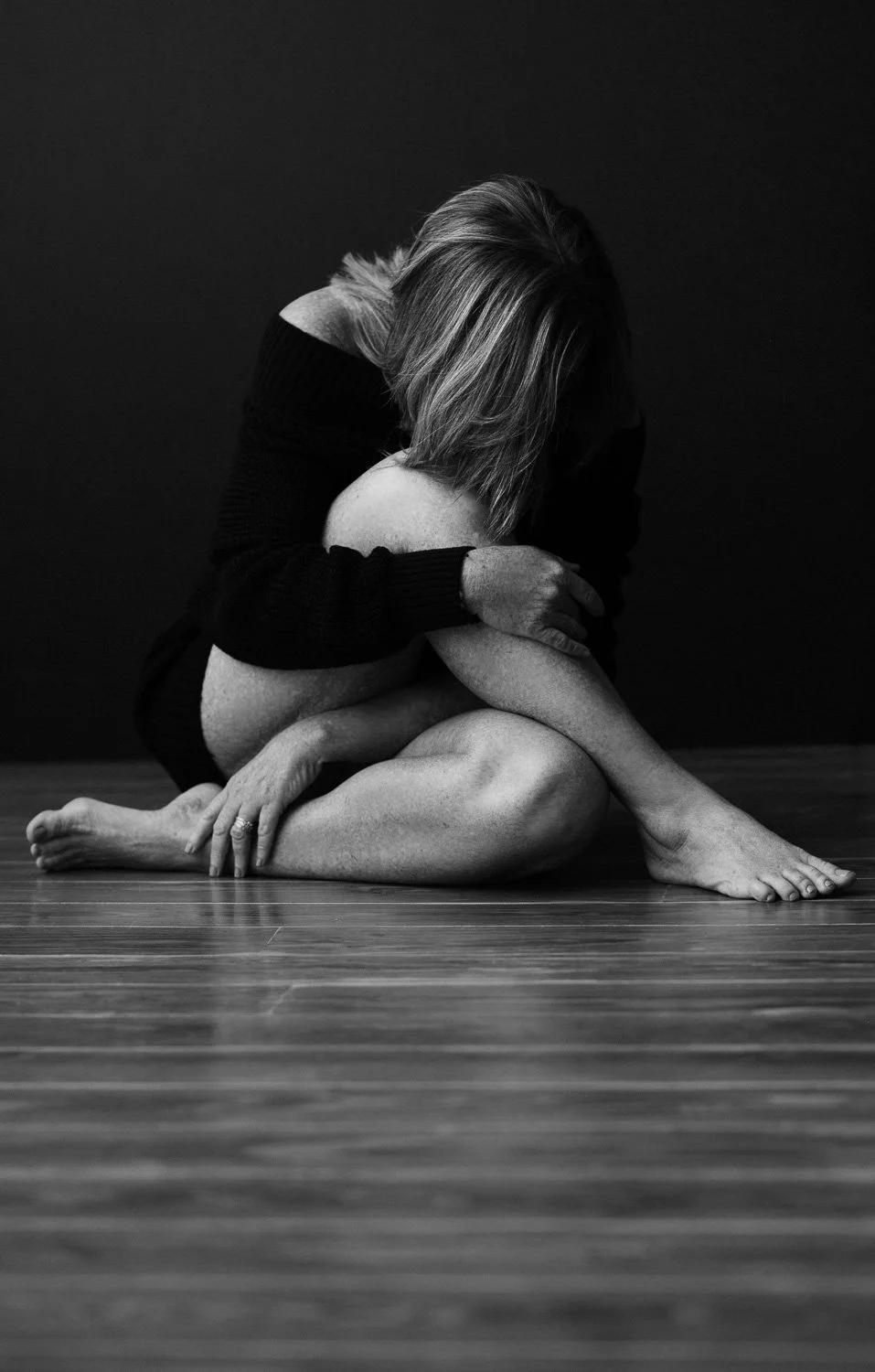 A woman sitting on a wooden floor with her knees drawn up to her chest, embracing her legs with her arms, and her head bowed down. She is wearing a black off-shoulder sweater with long sleeves. The photo is in black and white, with a dark background.