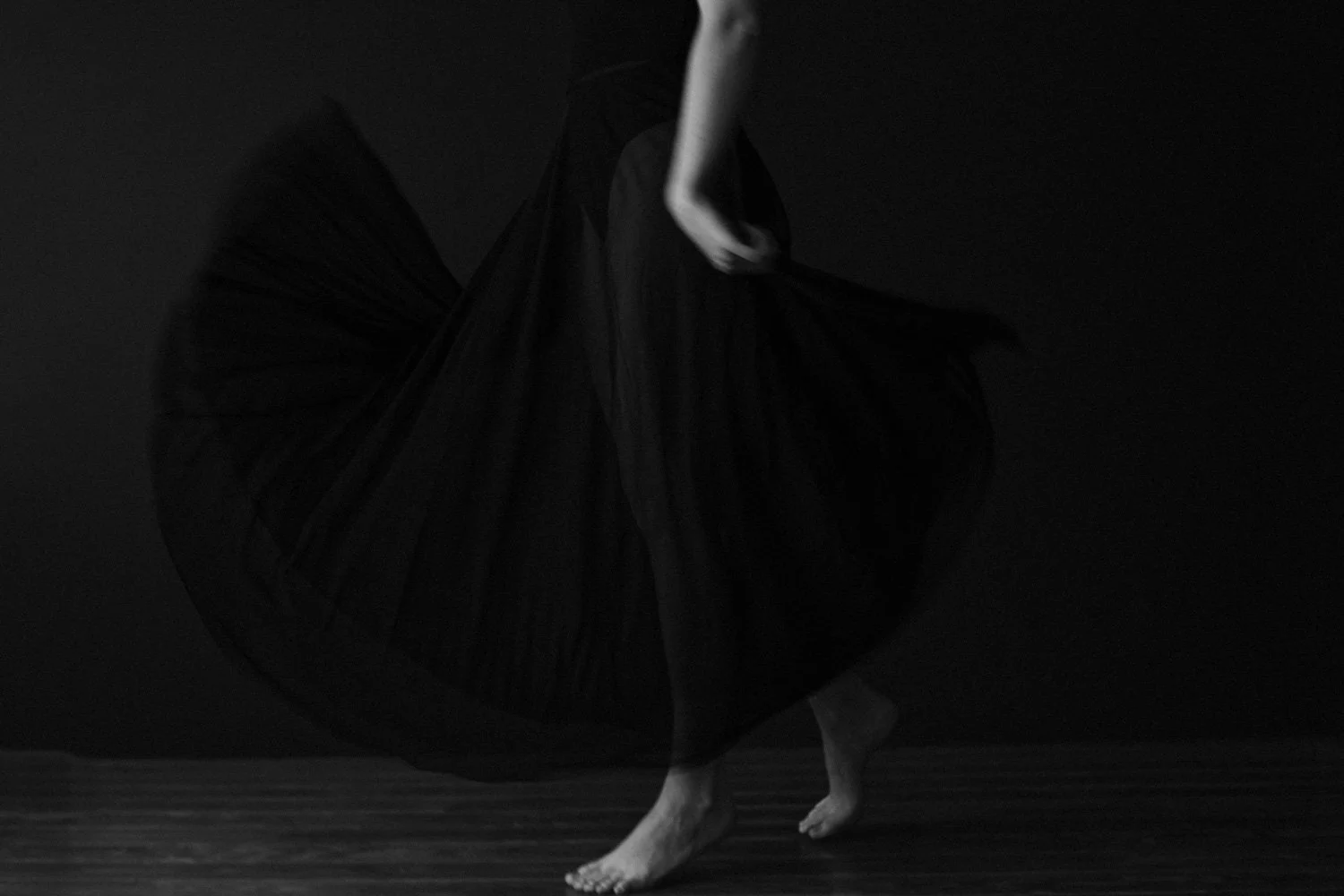 A person dancing barefoot in a long, flowing black skirt against a dark background.