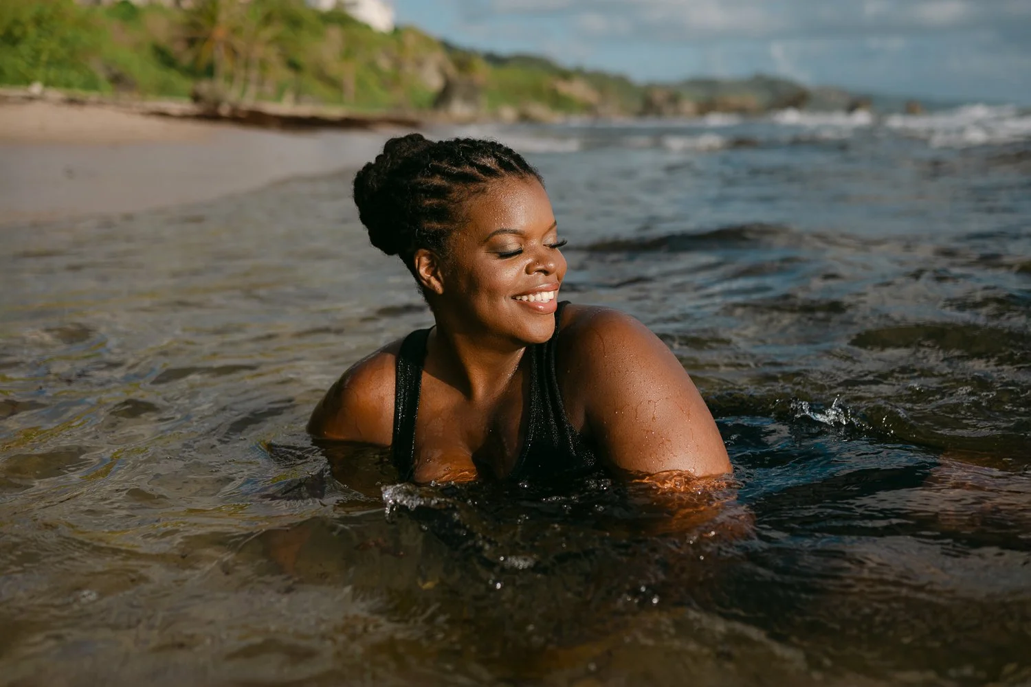 A woman with braided hair smiling and enjoying herself in the water at the beach, with a sandy shoreline and greenery in the background.