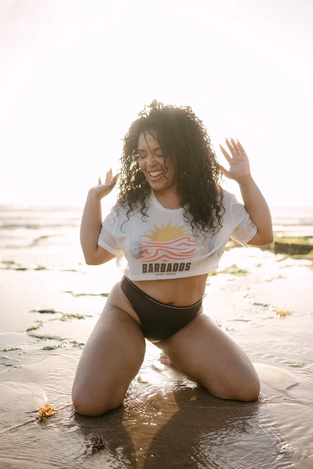 A woman with curly hair kneeling on a beach, smiling and enjoying the sun, wearing a Barbados t-shirt and black shorts.