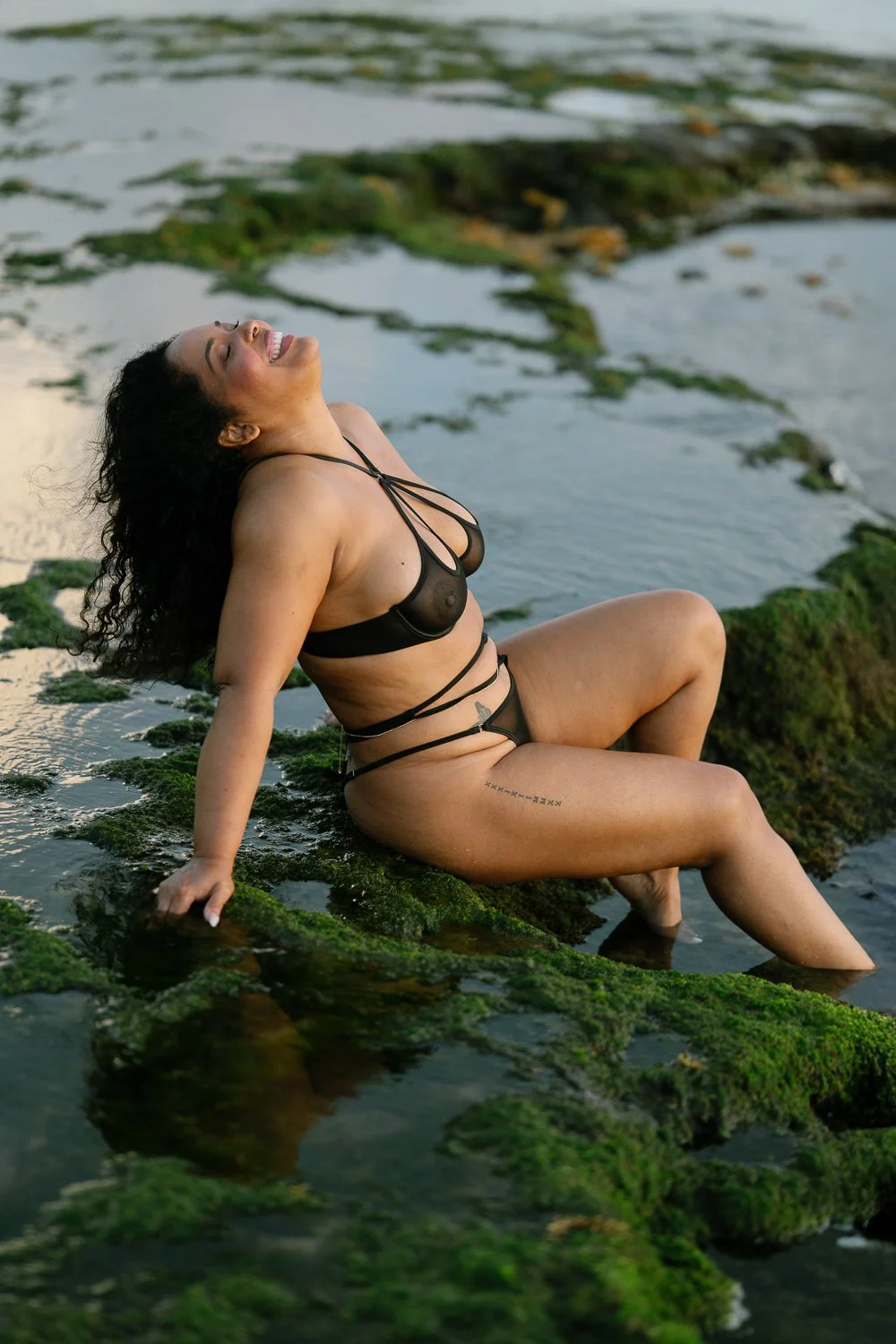 Woman in black lingerie sitting on moss-covered rocks by the water, smiling with eyes closed, enjoying the outdoors.