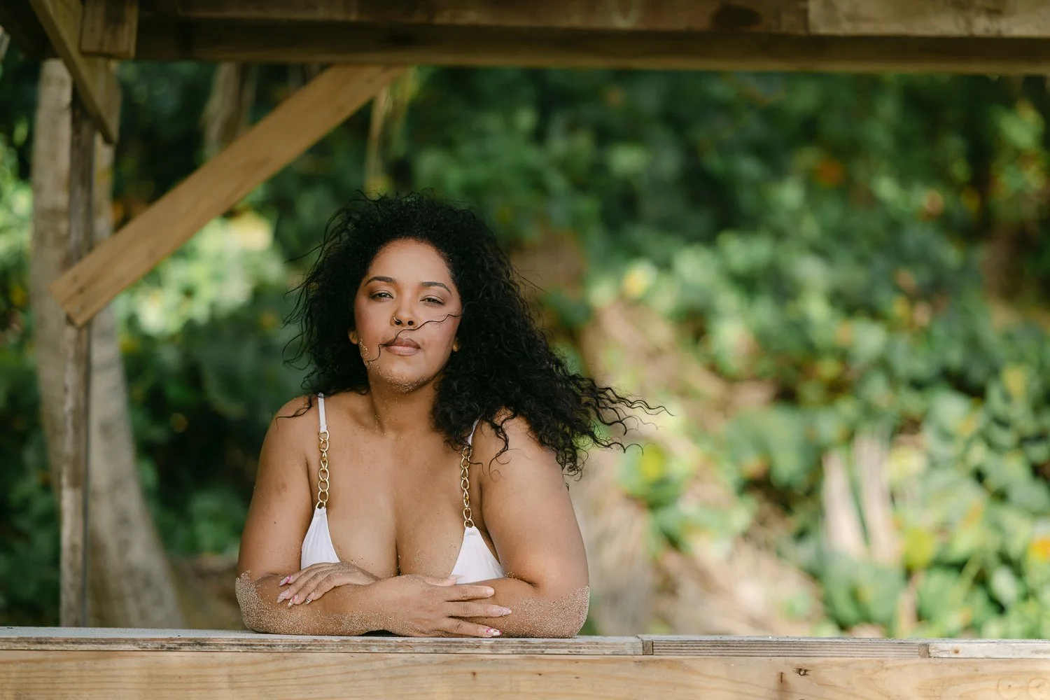 A woman with curly black hair and a nose ring sitting at a sandy beach, leaning on a wooden railing under a wooden structure with green foliage in the background.