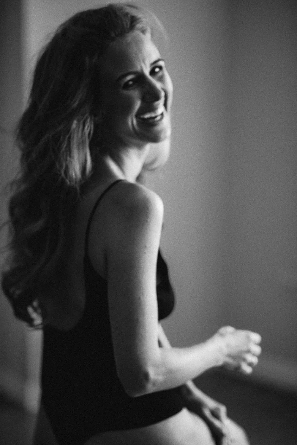 Black and white photo of a woman smiling, with long wavy hair, wearing a sleeveless top, in a relaxed pose.